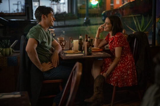Man in a green shirt and a woman in a red dress sit across from each other at a dimly lit restaurant table, smiling and talking.