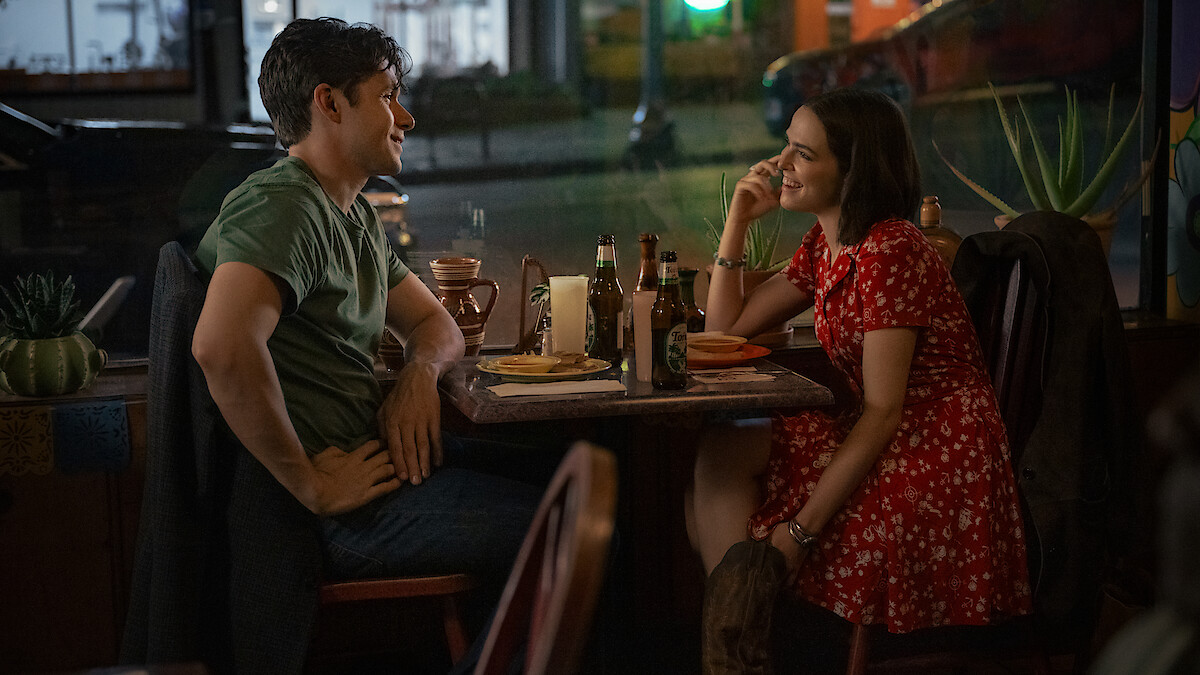 A man in a green shirt and a woman in a red floral dress sit across from each other at a small, dimly lit restaurant table, smiling while talking with drinks and plates between them on the table.