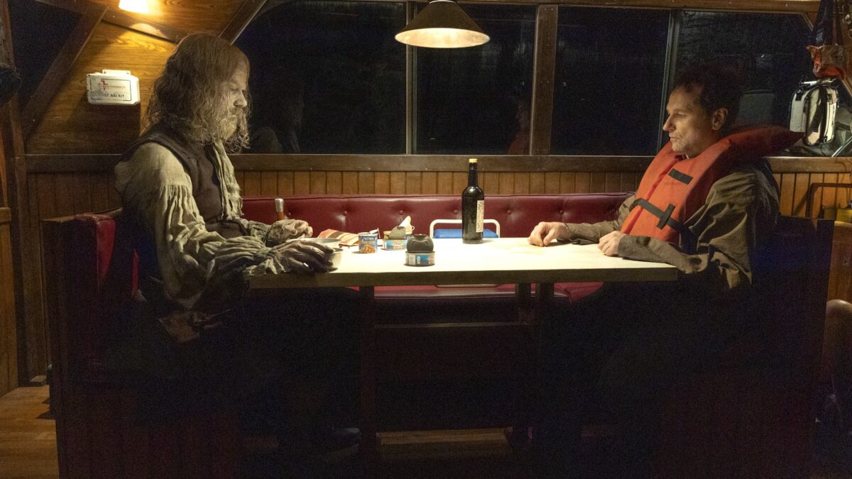 Two men sit across from each other in a wood-paneled booth at night; one wears a life jacket and a bottle of wine sits on the table.