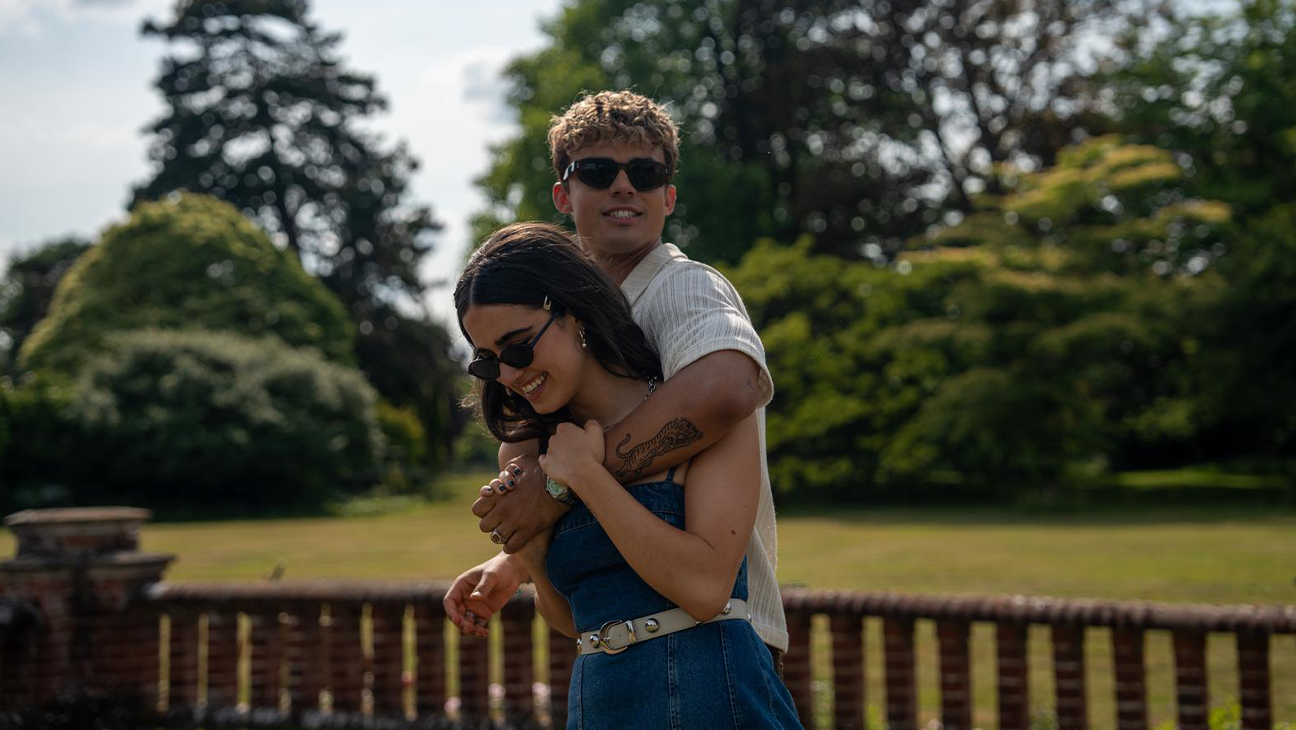 Couple in a park: man hugs woman from behind as they both wear sunglasses and smile happily in a sunny garden setting.