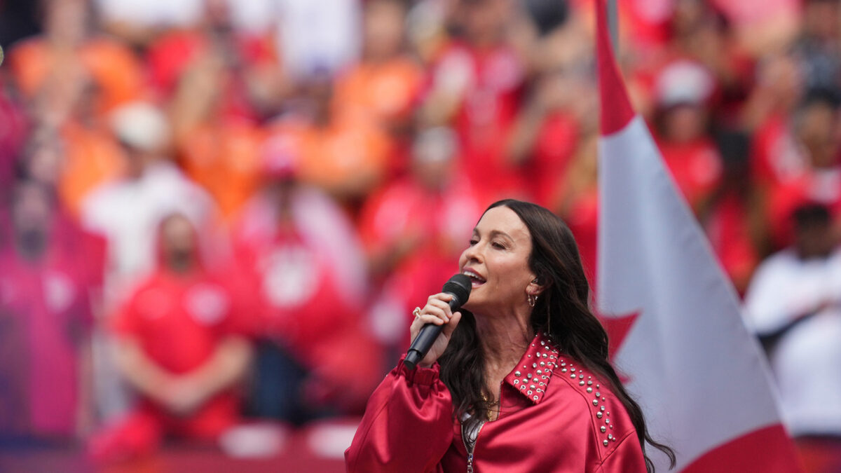 Female singer in a red studded jacket sings into a microphone on stage with a blurred red-clad crowd in the background.