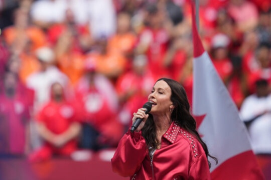 Female singer in a red studded jacket sings into a microphone on stage with a blurred red-clad crowd in the background.