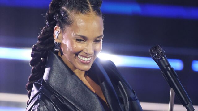 Smiling female singer with braided hair on stage, wearing an in-ear monitor and a black jacket by the microphone.