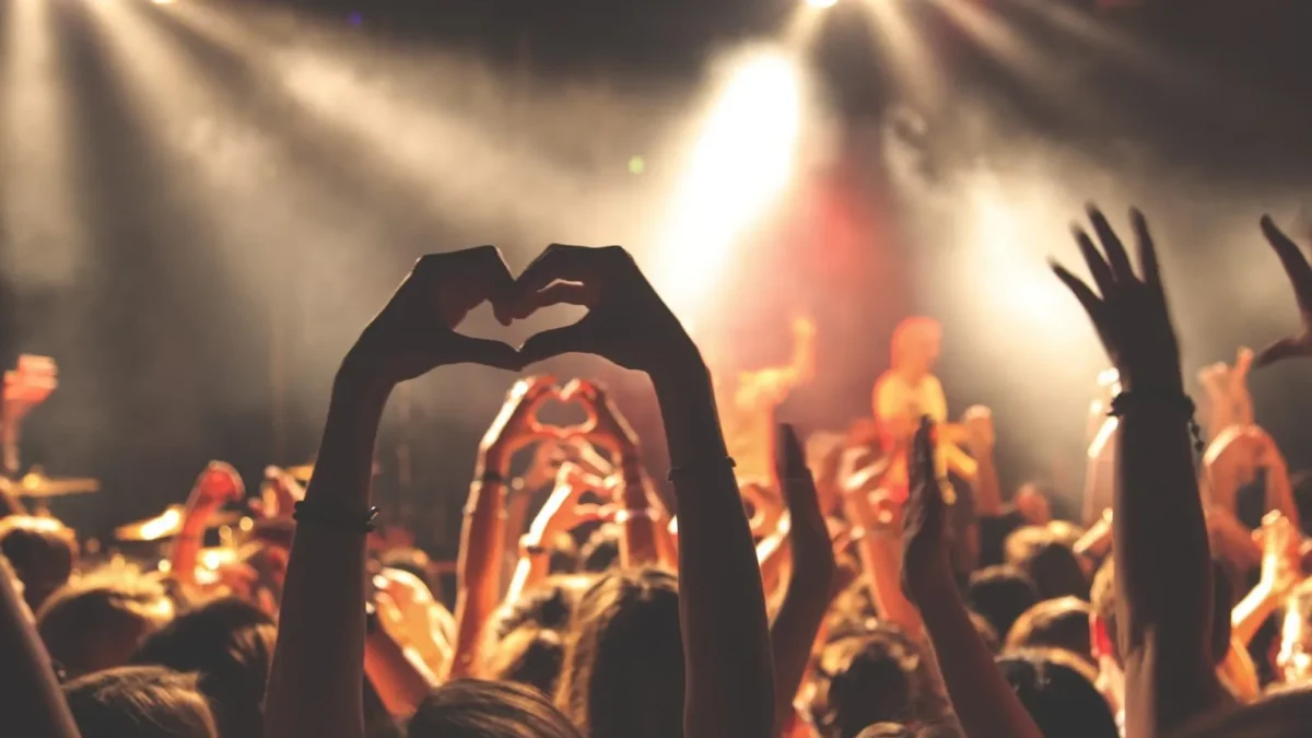 Crowd at a concert with hands forming a heart shape against bright stage lights.