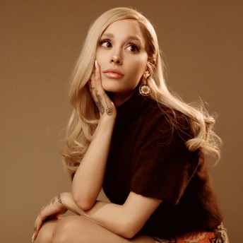 Portrait of a blonde woman in a dark top, resting her chin on her hand, with visible arm tattoos, against a warm beige background.