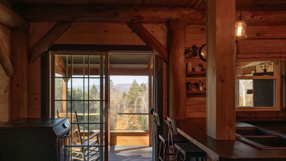 Rustic cabin interior with exposed log beams and a sliding glass door opening to a deck with wooden rocking chairs and a forested mountain view.