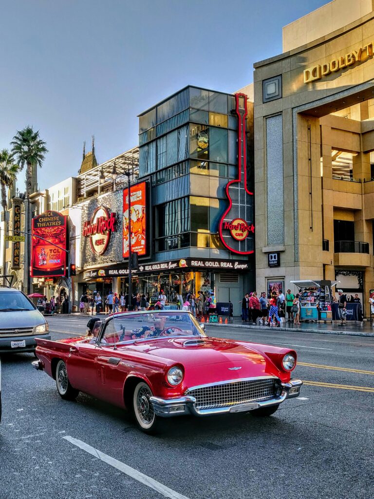 Red vintage convertible cruising down a sunny Hollywood street with the Hard Rock Cafe guitar sign and Dolby Theatre in the background.