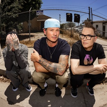 Three young men crouching on a sidewalk in front of a chain-link fence; left person covers face, middle man wears a light blue cap and has tattoos, right man wears glasses.