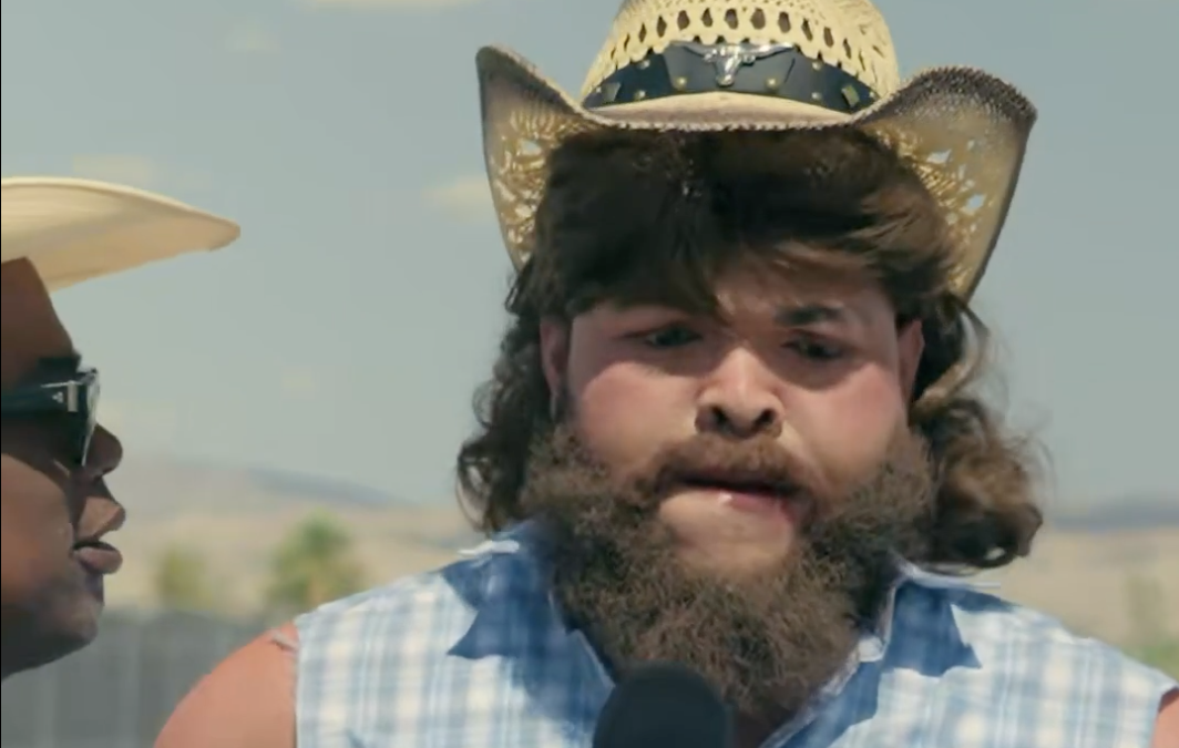Man with a large beard and straw cowboy hat speaks into a microphone during an outdoor interview.