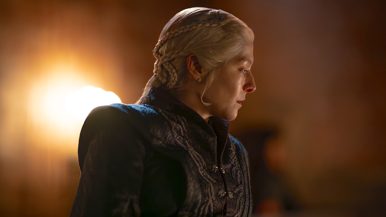 Profile of a blonde woman with intricate braids, wearing a dark ornate jacket, lit by warm backlight.