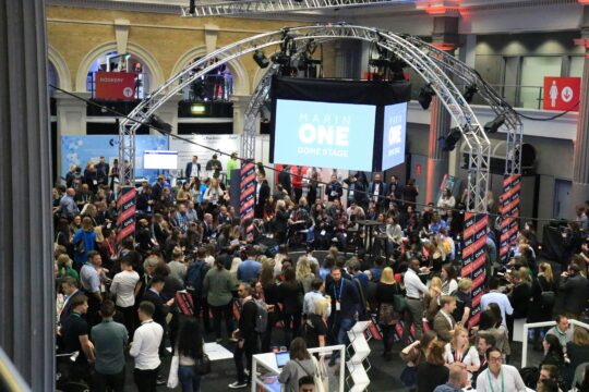 Busy conference hall with a large suspended stage screen reading 'Marin One Dome Stage' surrounded by truss arches and a crowd of attendees.