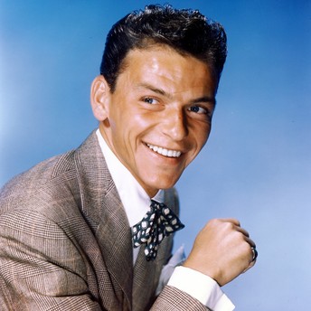 Portrait of a smiling man in a brown checkered suit and bow tie against a blue studio backdrop.