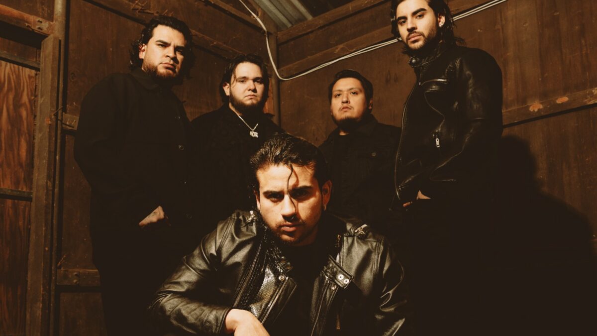 Five men in dark clothing pose in a dim, wood-paneled space; the foreground man wears a black leather jacket and looks at the camera.”] ,