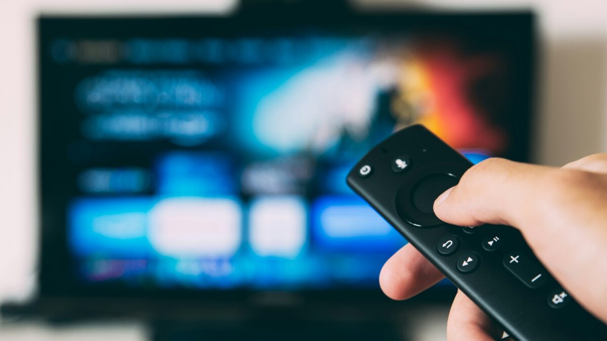 Close-up of a hand holding a black remote control with blurred TV screen in the background, showing playback buttons.