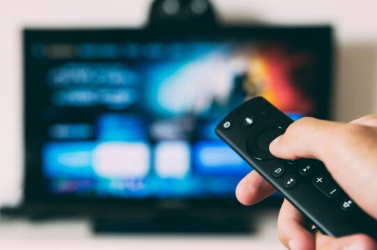 Close-up of a hand holding a black remote control with blurred TV screen in the background, showing playback buttons.