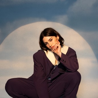 Woman in a dark purple suit with a white lapel crouches, resting her chin on her hand, against a cloudy blue backdrop with a large circular light behind her.