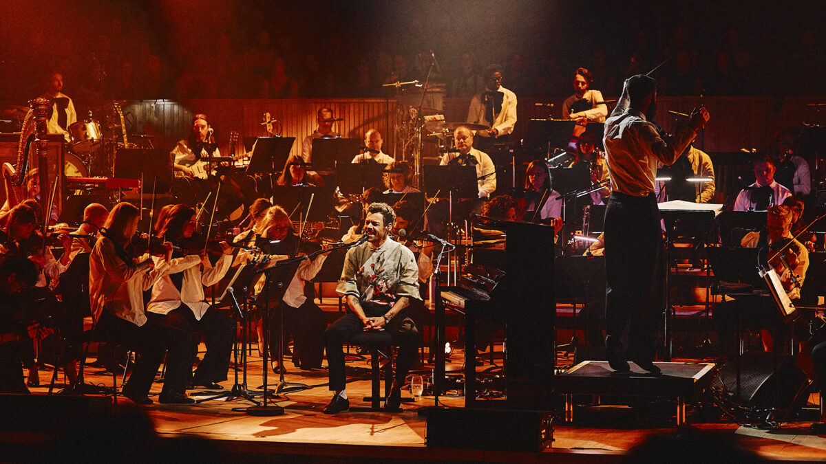 Conductor on a raised podium directs a full orchestra on stage, while a singer sits center front with a microphone.