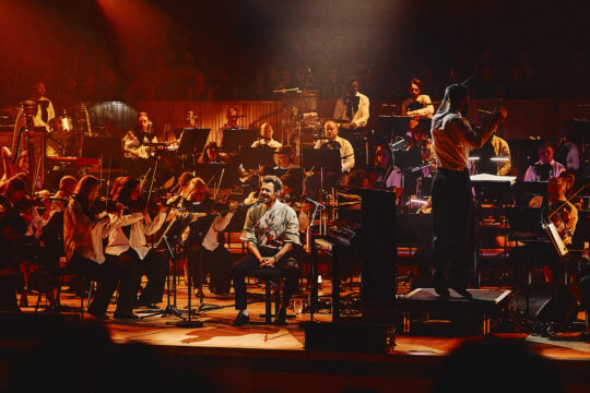 Conductor on a raised podium directs a full orchestra on stage, while a singer sits center front with a microphone.
