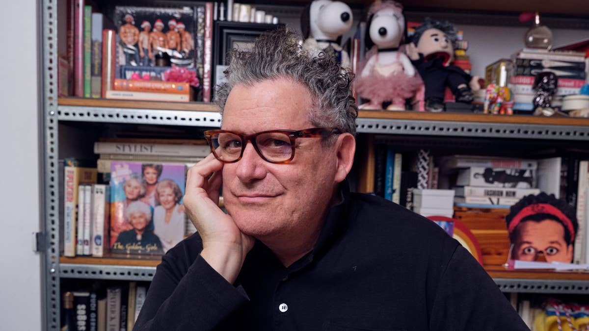 Portrait of a middle-aged man with brown glasses resting his chin on his hand, in front of a bookshelf filled with books and toys