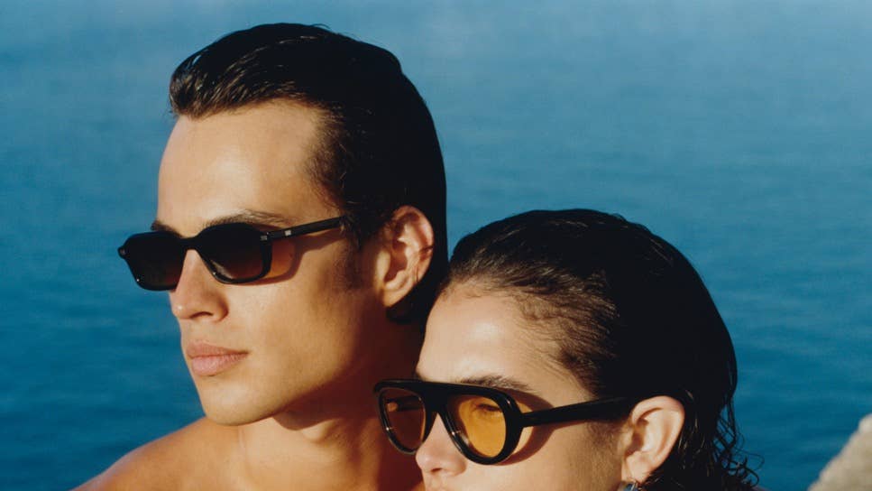 Two people wearing sunglasses stand side by side by a blue ocean sea horizon behind them.