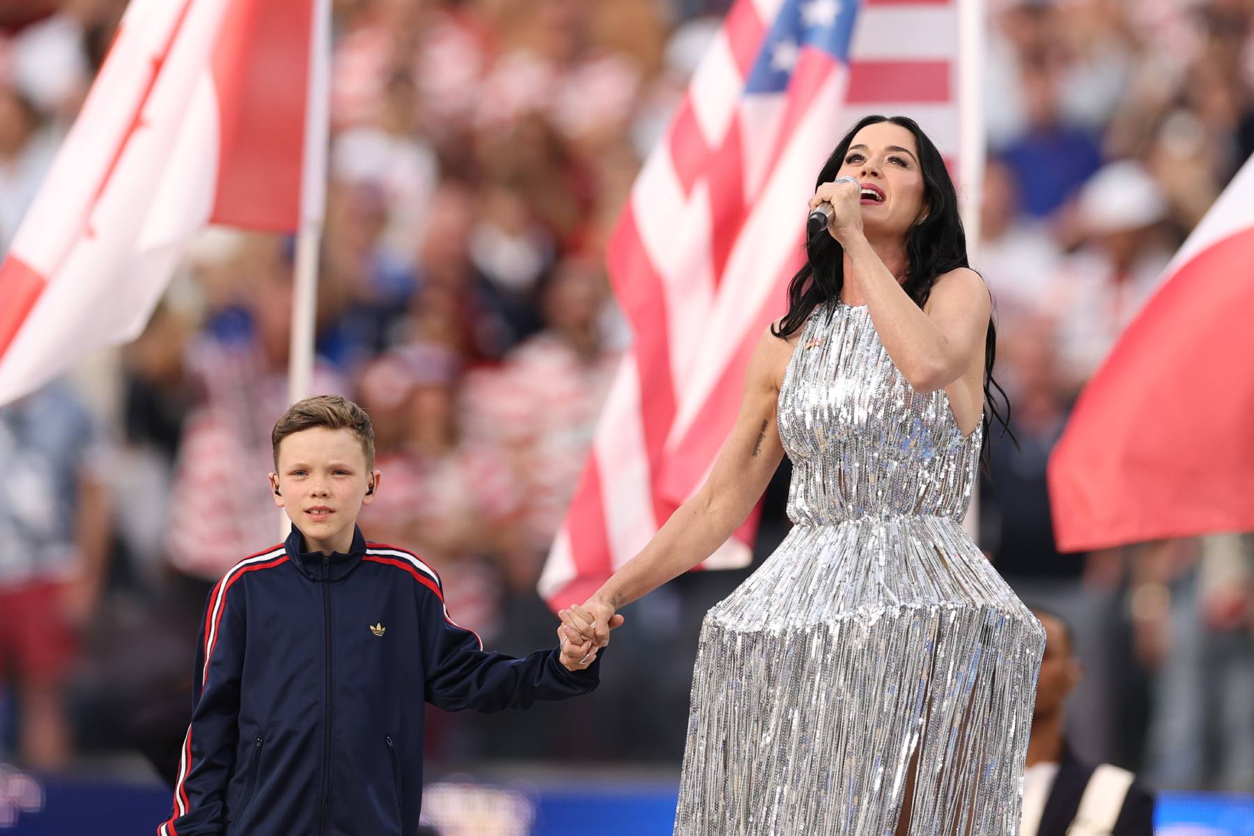 Singer in a silver glitter dress performs on stage while holding hands with a boy in a navy jacket; waving flags in the background.