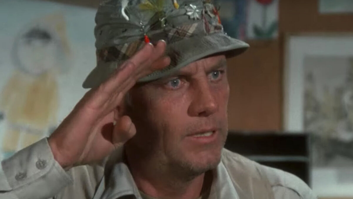 Man in a camo hat decorated with flowers salutes, raising his right hand to his forehead indoors.