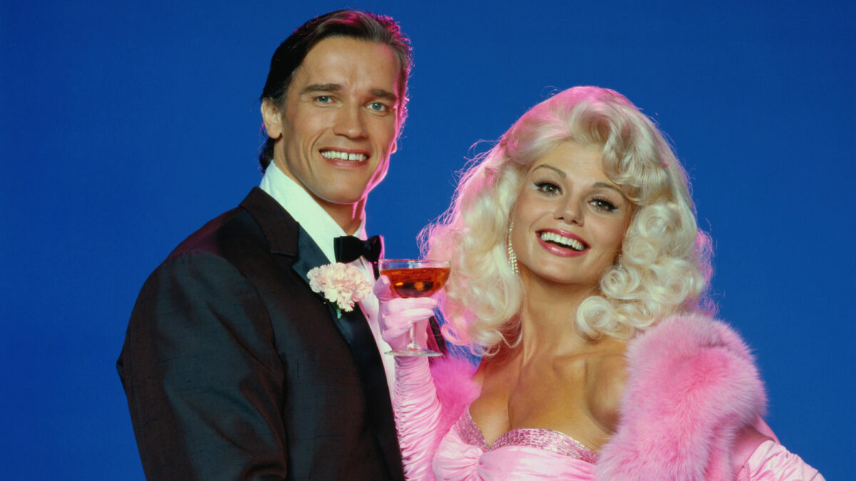 Smiling couple in formal attire against a blue backdrop; man in a tuxedo, woman in a pink gown with a fur stole, holding a champagne glass.