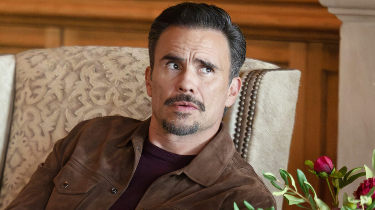 Man in a brown jacket sits in a patterned upholstered chair, looking to the side with a surprised/concerned expression, flowers nearby on the right.