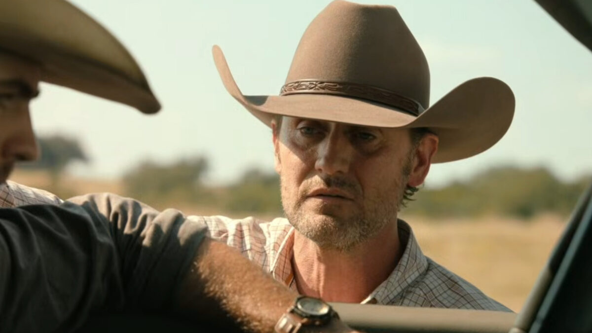 Man wearing a brown cowboy hat leans toward a car window, speaking to a companion off-screen in a desert-like setting.