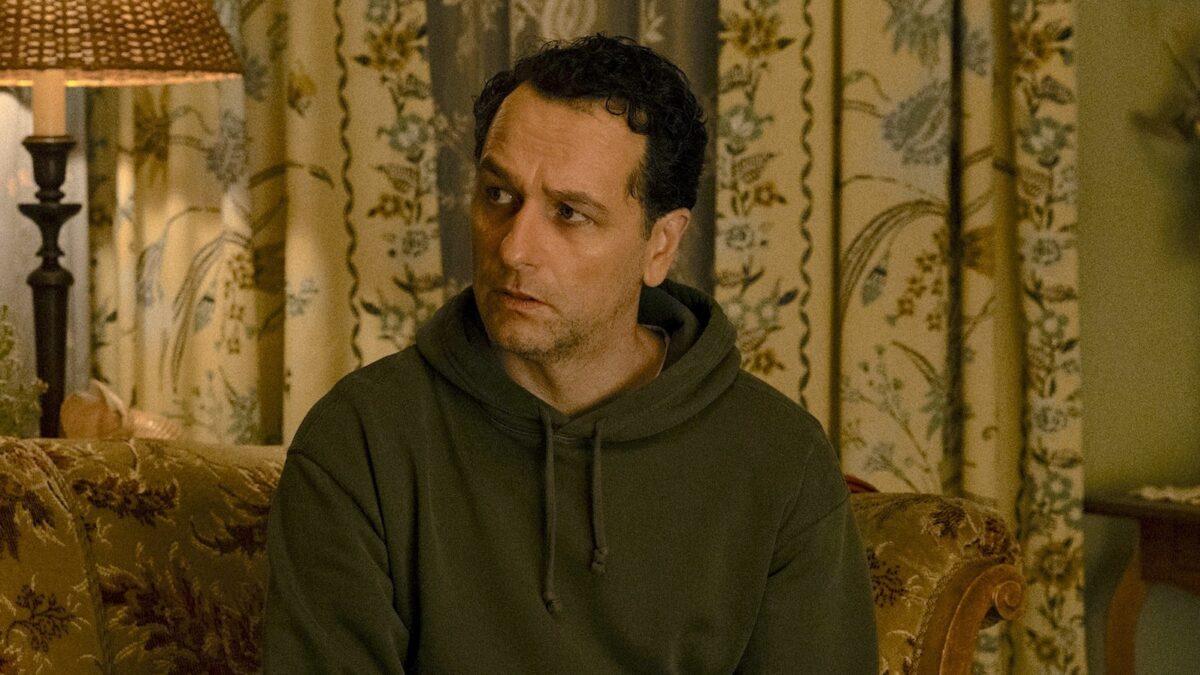 Man in a dark hoodie sits on a patterned sofa in a cozy living room, looking to the right.