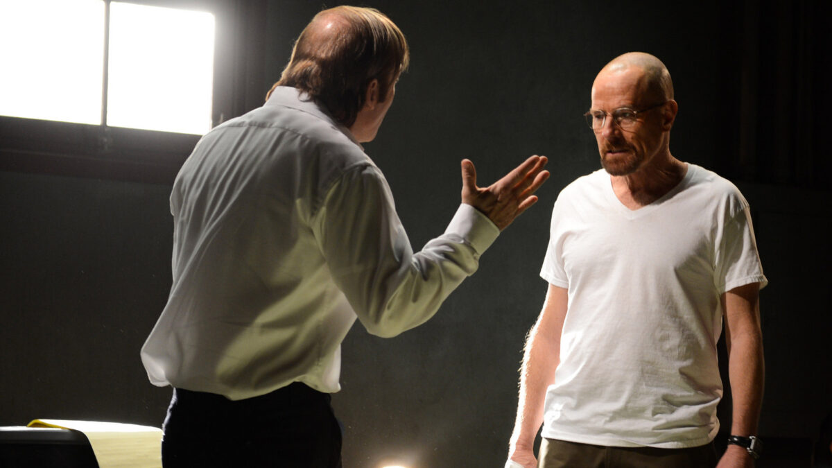 Two men in a dim rehearsal space; one facing the other with an outstretched hand as if explaining something