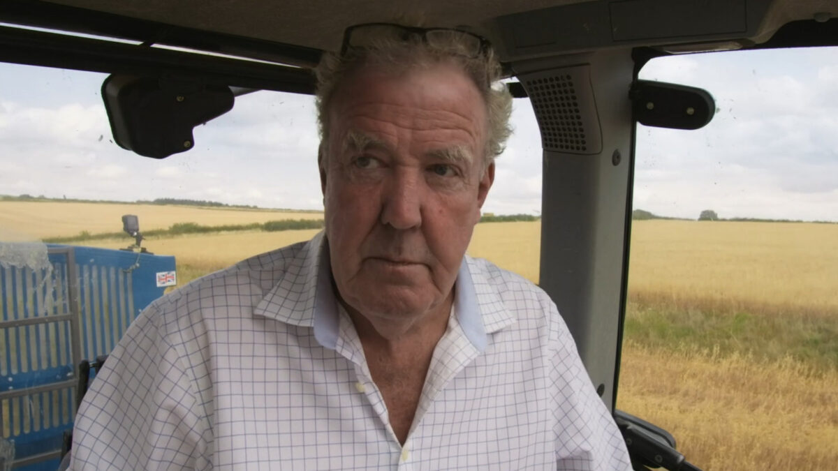 Older man in a white checkered shirt sits in a tractor cab with a golden field outside.