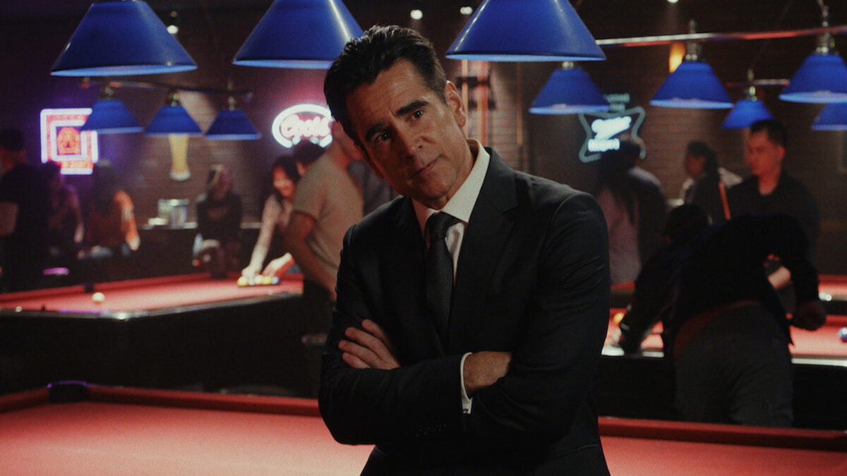 Man in a dark suit stands with arms crossed in a dimly lit billiards hall, blue lamps overhead.