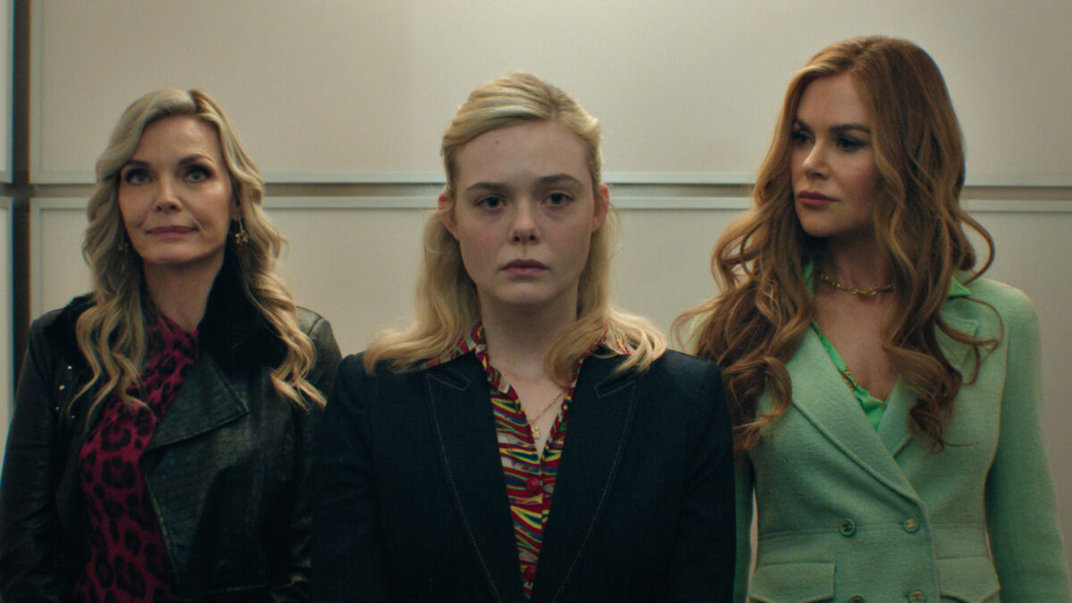 Three women stand side by side, looking straight ahead in an elevator; left wears a black leather jacket and red leopard print top, center in a dark blazer with a colorful patterned shirt, right in a mint green coat with a gold necklace.