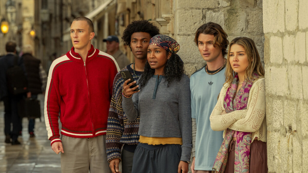 Five diverse friends stand along a stone wall, looking intently at something off to the right.