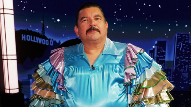 Smiling man in a bright turquoise ruffled costume on a TV studio set with a Hollywood backdrop.
