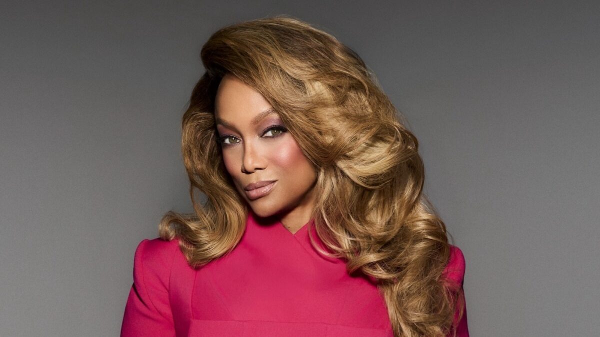 Portrait of a woman with voluminous blonde hair wearing a bright pink top, looking at the camera against a gray background.