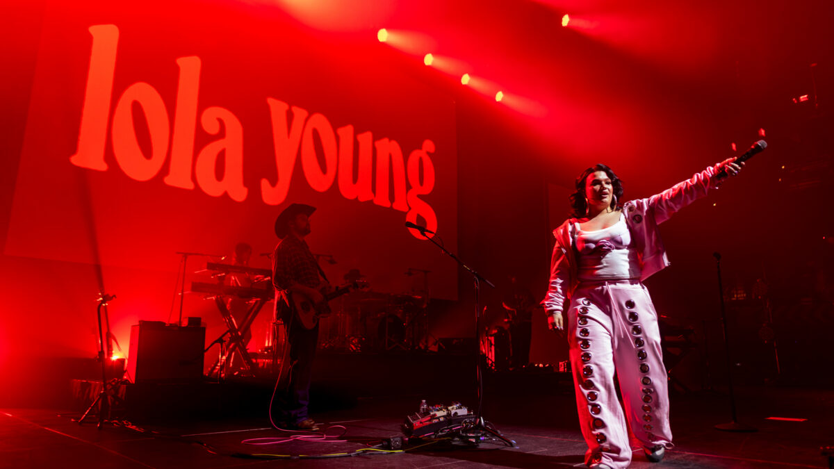 Female singer on stage with microphone, red lighting, band in background, and 'Lola Young' text on screen behind.