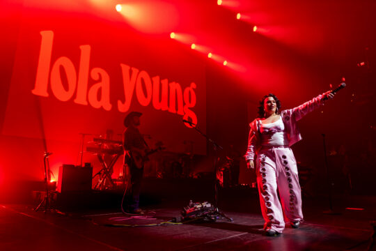 Female singer on stage with microphone, red lighting, band in background, and 'Lola Young' text on screen behind.