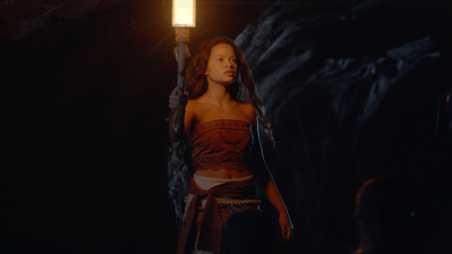 Woman in a red patterned wrap holding a lit torch in a dark cave, face softly illuminated by the flame.