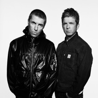 Two men pose side by side in black-and-white; left man wears a shiny puffer jacket, right man in a dark button-down shirt with serious expressions.