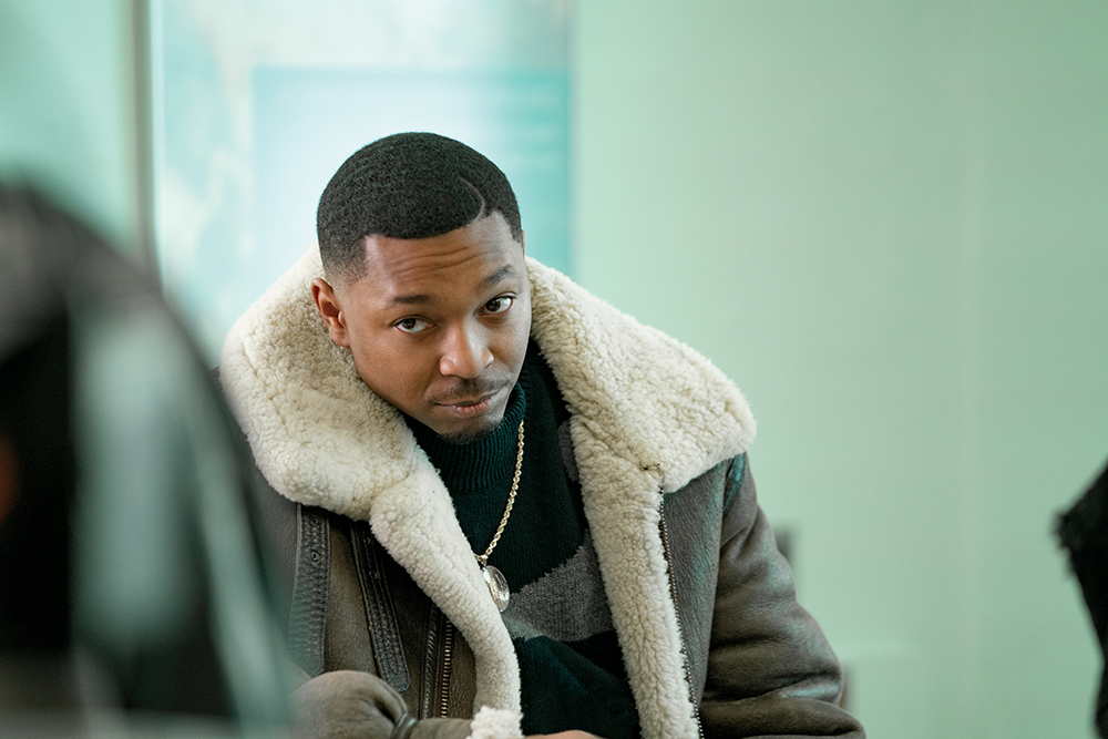 Young man wearing a brown shearling-lined coat and gold necklace looks at the camera in a pale green room.