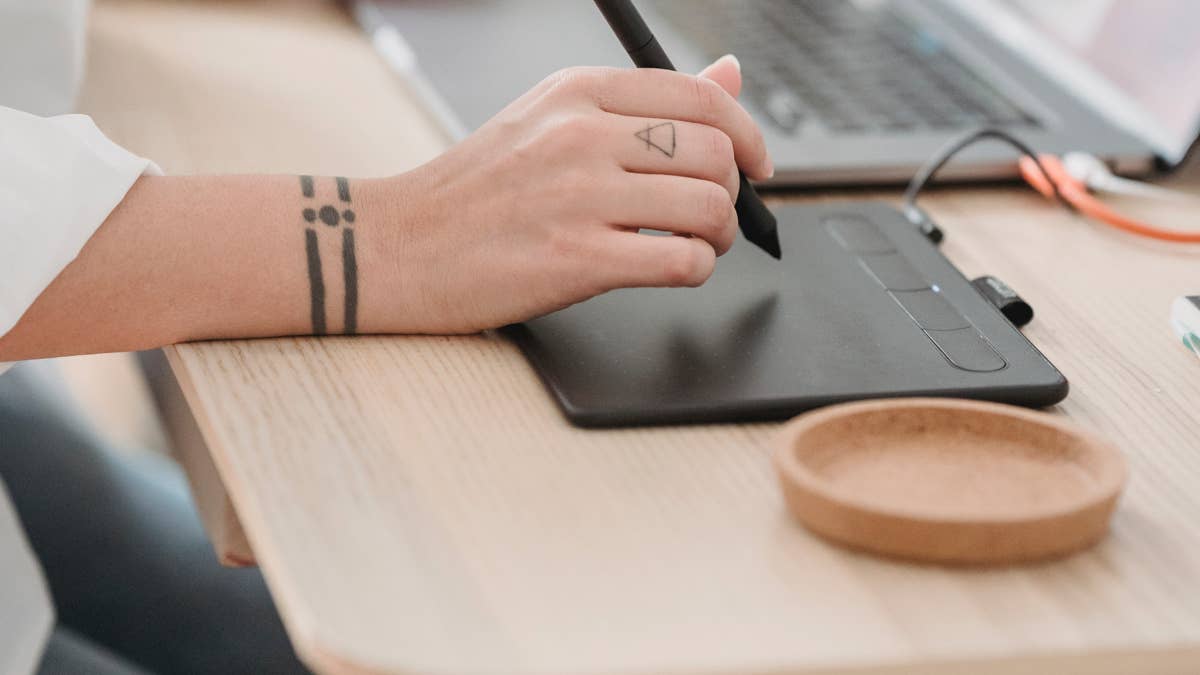 Person drawing on a graphics tablet with a stylus at a wooden desk, a cork coaster nearby.