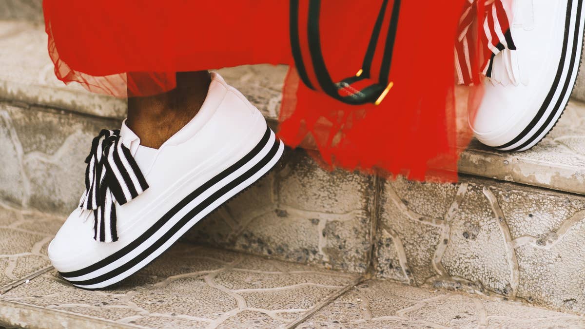Close-up of white platform sneakers with black stripes and bow laces, worn with a red dress on stone steps.