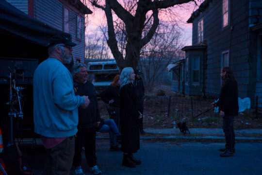 People stand outside at dusk in a blue-tinged scene, talking near a small dog on a leash with houses and a large tree in the background.