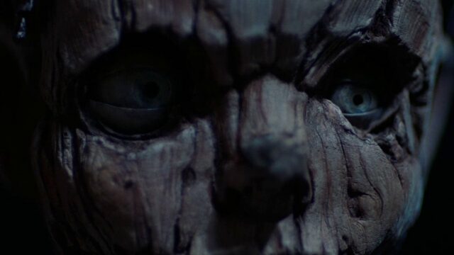 Close-up of a cracked, weathered face with piercing blue eyes in a dark setting.