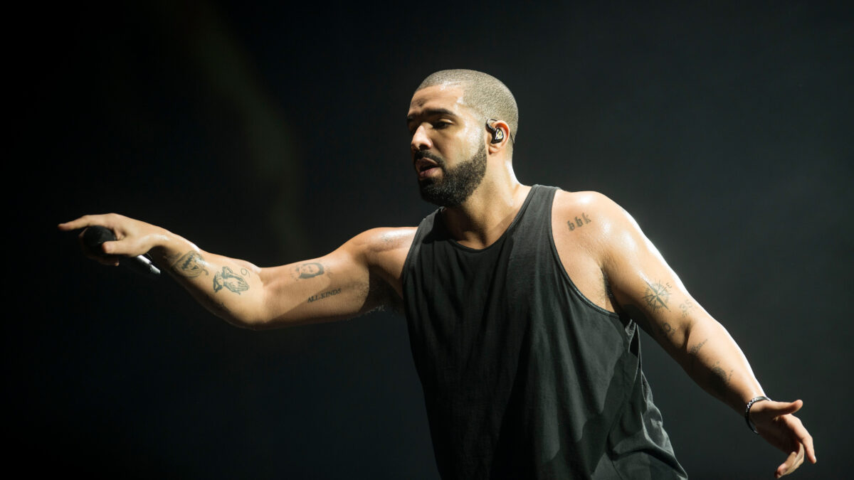 Male performer on stage in a black sleeveless shirt, mic in hand, arms outstretched against a dark background.