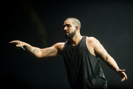 Male performer on stage in a black sleeveless shirt, mic in hand, arms outstretched against a dark background.