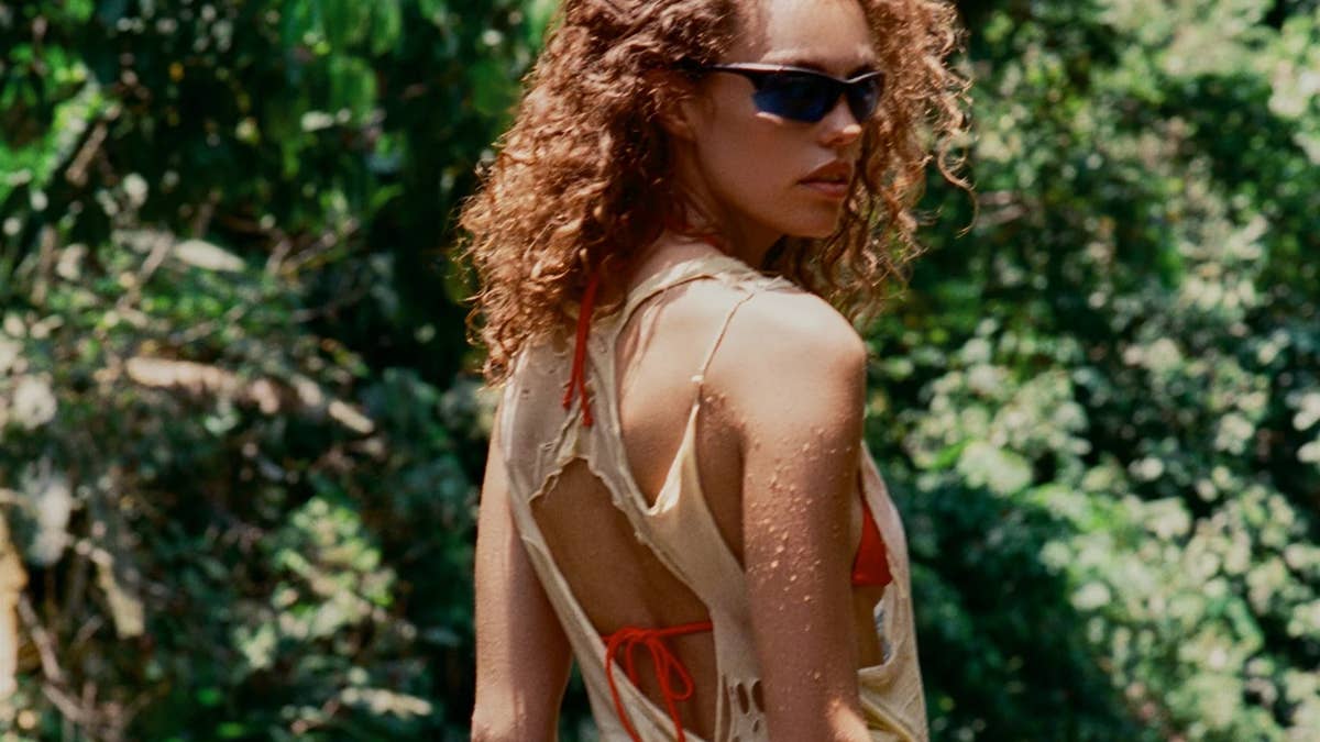 Woman with curly hair wearing sunglasses and a torn beige top in a sunlit green outdoor setting