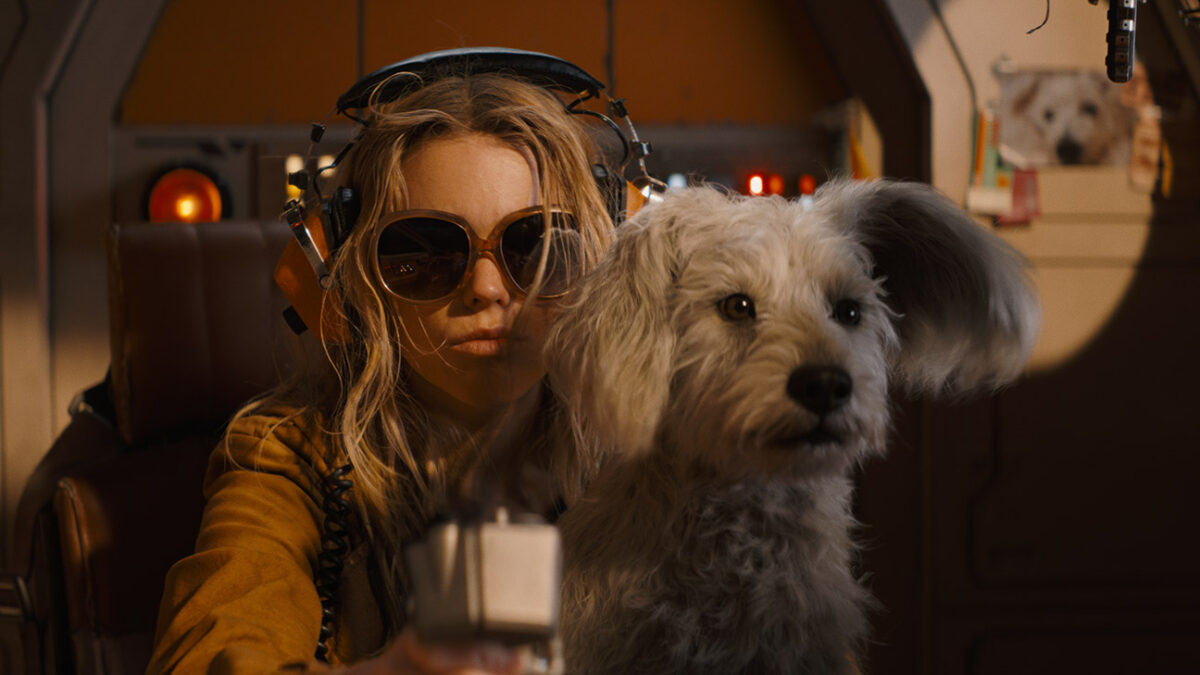 Woman wearing large sunglasses and headgear in a spaceship cockpit with a fluffy dog beside her, holding a control or device toward the camera.